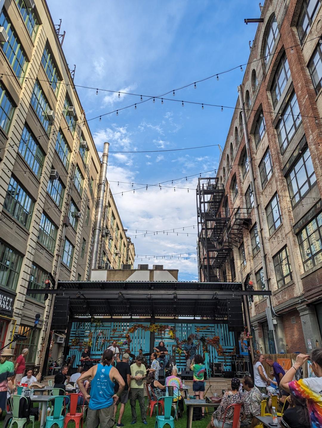 A local band on a small stage in brooklyn with a small crowd. The stage is between two building, like a courtyard