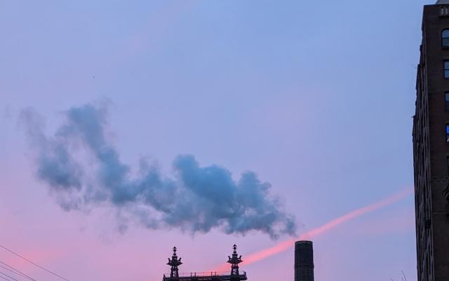 A photograph of the Queensboro Bridge in NYC at Sunrise. The sky is very pink and purple. The bridge is big. A smoke stack is blowing steam. 